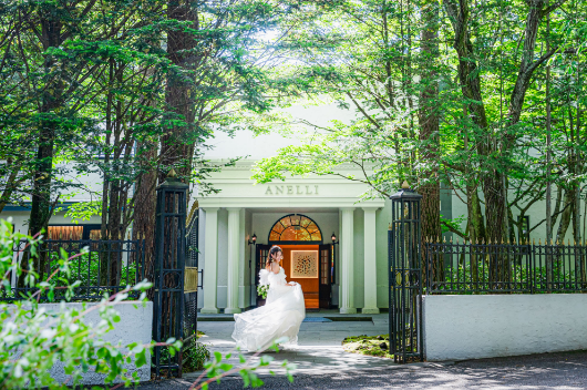 【アネーリ軽井沢の結婚式】軽井沢の豊かな緑に囲まれた結婚式場の外観と、ウェディングドレスの花嫁。森の中に佇む白亜の会場入口前で、自然光を浴びながら撮影するロケーションフォト。軽井沢でのガーデンウェディングやリゾートウェディング、前撮りをお考えの新郎新婦におすすめ。
