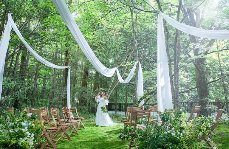 アネーリ軽井沢の森のウェディング会場。白い布で装飾された祭壇に立つ花嫁。緑の芝生と木製の椅子が並ぶ、軽井沢リゾートウェディングの自然な挙式スタイル。