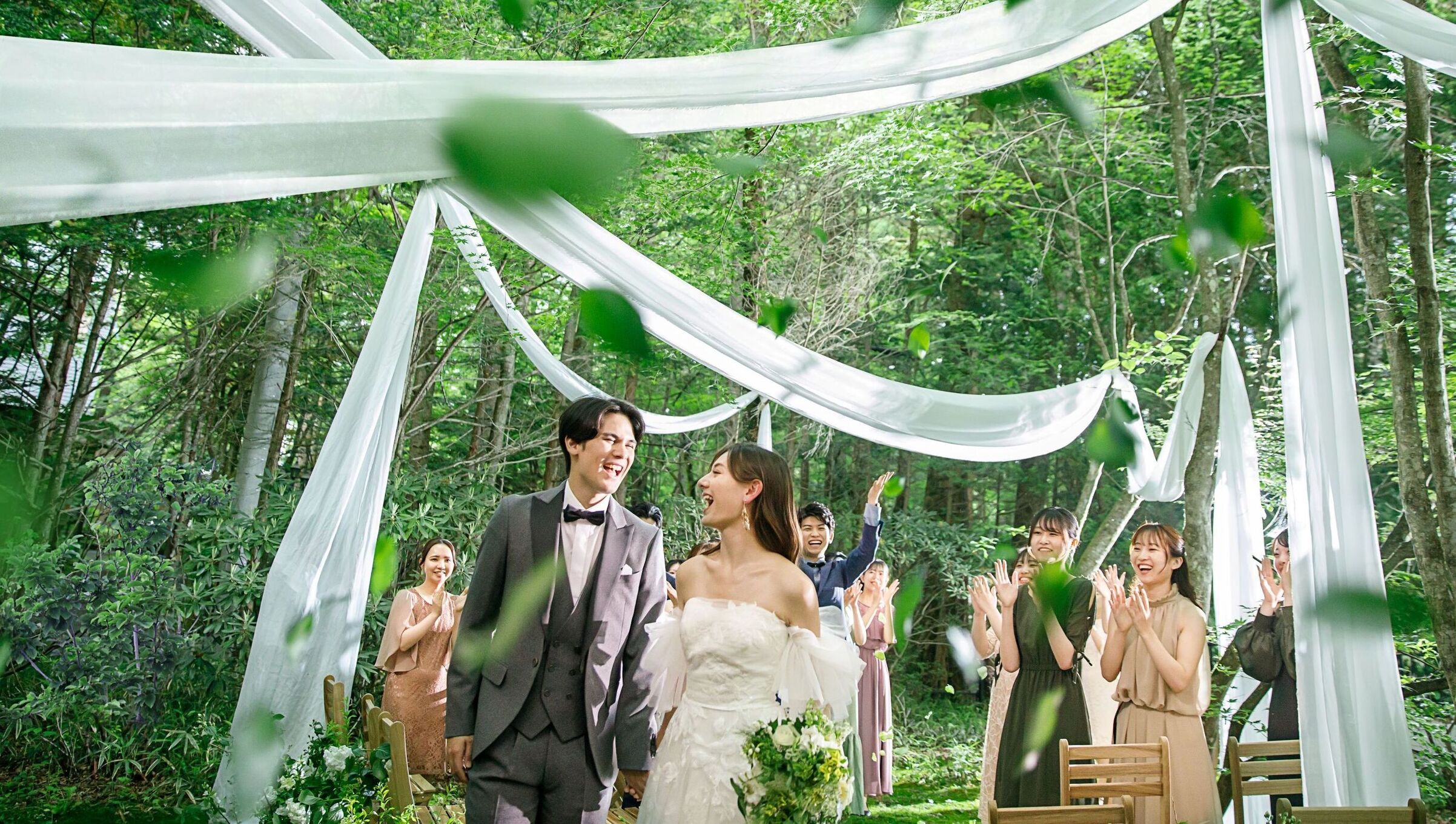 【軽井沢の自然に囲まれた森のウェディング】 新郎新婦が緑の木漏れ日の中を歩くガーデン挙式。アネーリ軽井沢で叶える、笑顔あふれるロケーションフォト。