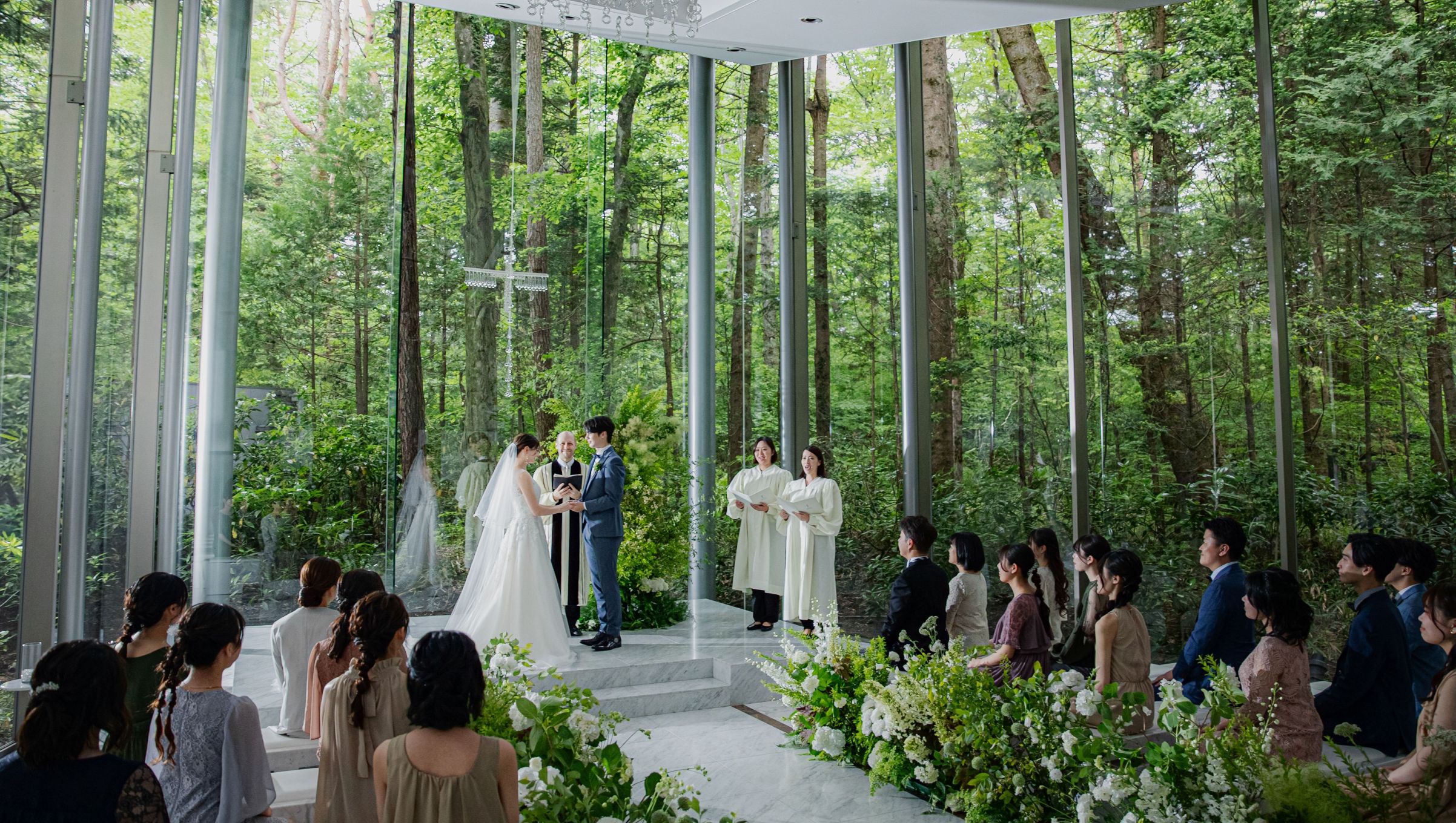 軽井沢のガラス張りチャペルで行われるウェディングセレモニー。森の木々の中で、聖歌隊の歌声とゲストの温かい眼差しに包まれながら愛を誓う新郎新婦。アネーリ軽井沢の挙式シーン。