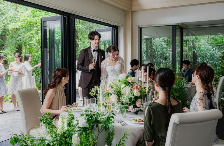 アネーリ軽井沢の結婚式披露宴会場。新郎新婦が軽井沢の自然に面したテラスから入場し、ゲストへ感謝を伝える様子。ブラウンのタキシードとレースのウェディングドレス。白と緑のナチュラルな装花がテーブルを彩る。ゲストハウスウェディングならではの親密でアットホームなリゾート婚のイメージ。