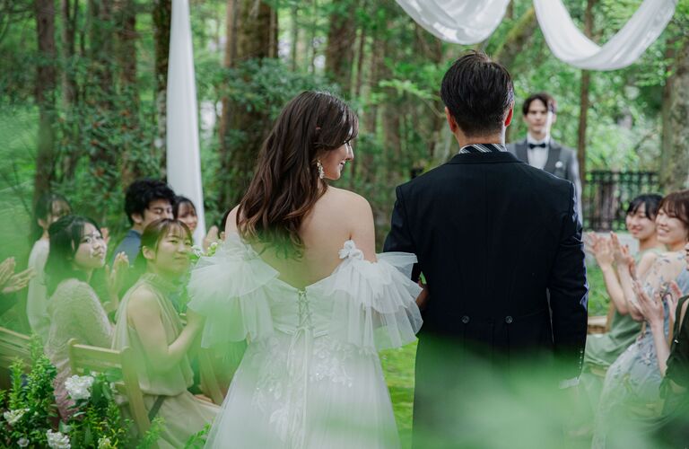 白いドレープが飾られたガーデン挙式会場。ゲストの拍手に迎えられ入場する新郎新婦。軽井沢の緑あふれるリゾートウェディング。