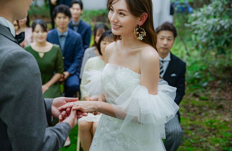 指輪交換をする新郎新婦。純白のオフショルダーウェディングドレスと自然派のイヤリングが印象的な、軽井沢の自然光が美しい挙式シーン。