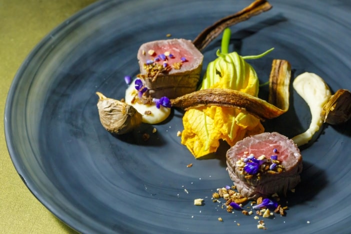 アネーリ軽井沢の信州フレンチを象徴するメインディッシュ。仔羊の低温調理と花ズッキーニ、黒ニンニクを洗練された器に芸術的に盛り付け。軽井沢ウェディングの美食による最上級のおもてなし。