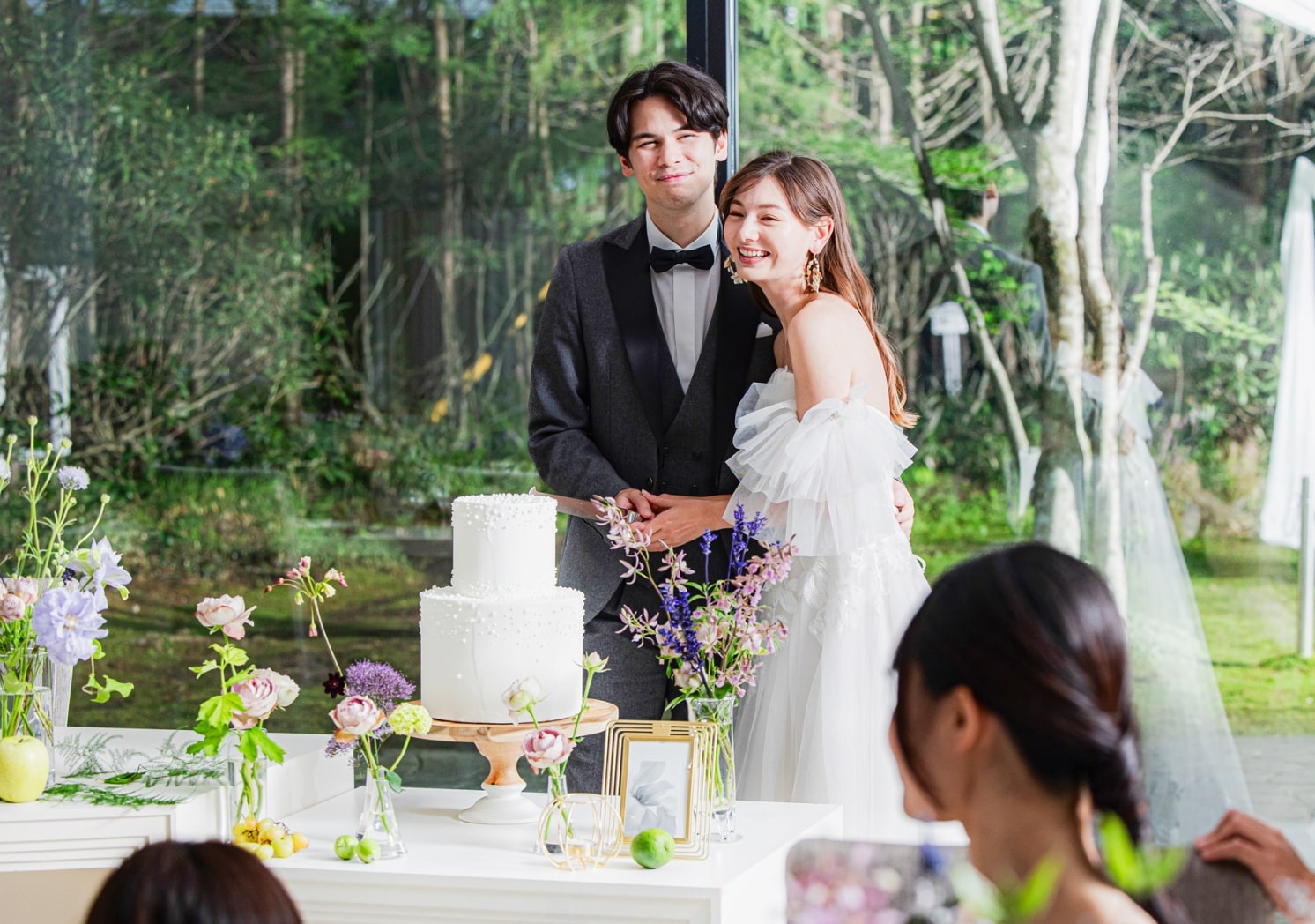 アネーリ軽井沢の全面ガラス張りの披露宴会場。軽井沢の緑豊かな森を背景にケーキカットを楽しむ新郎新婦。自然光溢れる貸切邸宅で叶えるリゾートウェディングの幸せな瞬間。