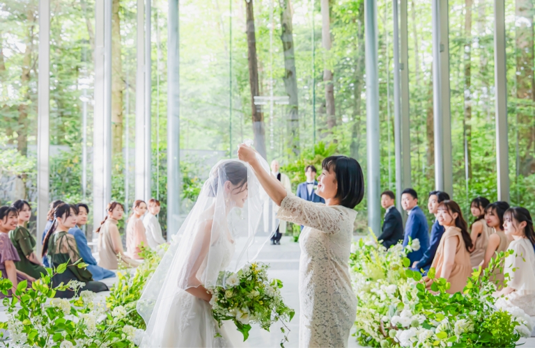 アネーリ軽井沢の森のチャペルでの挙式。母親の愛が伝わるベールダウンの儀式。ウェディングドレスの新婦とゲストを軽井沢の豊かな緑が祝福する、心温まるシーン。