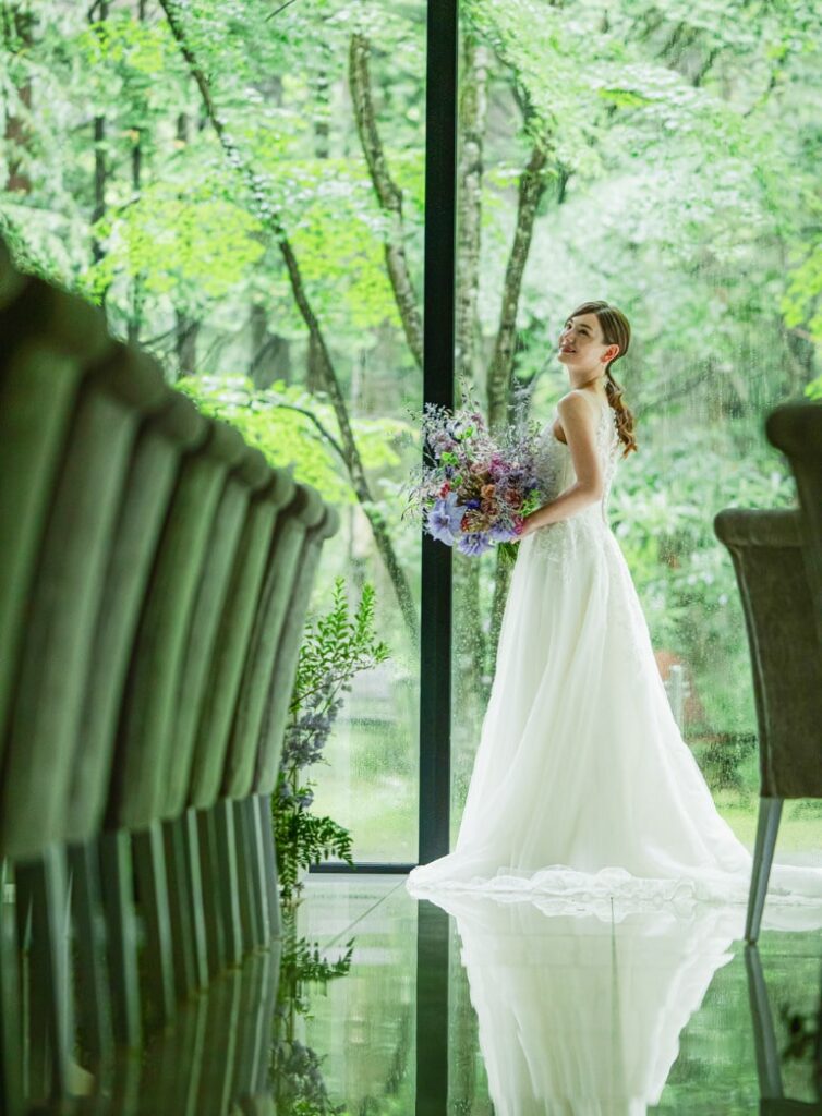 新緑の軽井沢の森と一体化する貸切の披露宴会場。大きな窓辺に立ち、ブーケを見つめ微笑む新婦のポートレート。森の緑がウェディングドレスを引き立てる、ナチュラルで感動的な瞬間。