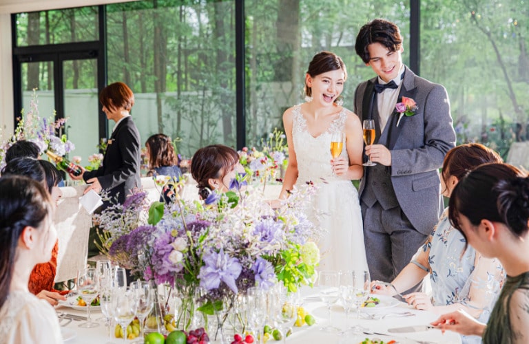 軽井沢の森に面した明るいパーティ会場。ゲスト一人ひとりと交流しながら、笑顔でテーブルを回る新郎新婦。ナチュラルでアットホームな雰囲気のウェディングパーティの様子。