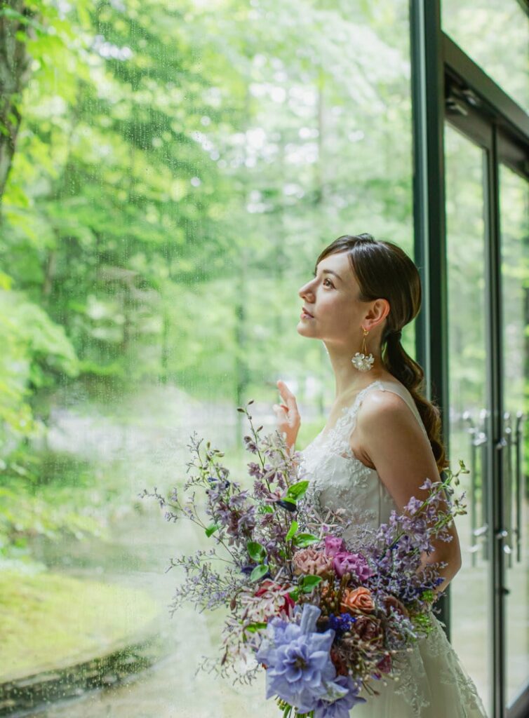 軽井沢の森の絶景を背景に、窓辺に立つウェディングドレス姿の新婦。自然光が美しく差し込む披露宴会場での、ブーケを持つ花嫁のポートレート。ナチュラルリゾートウェディング。