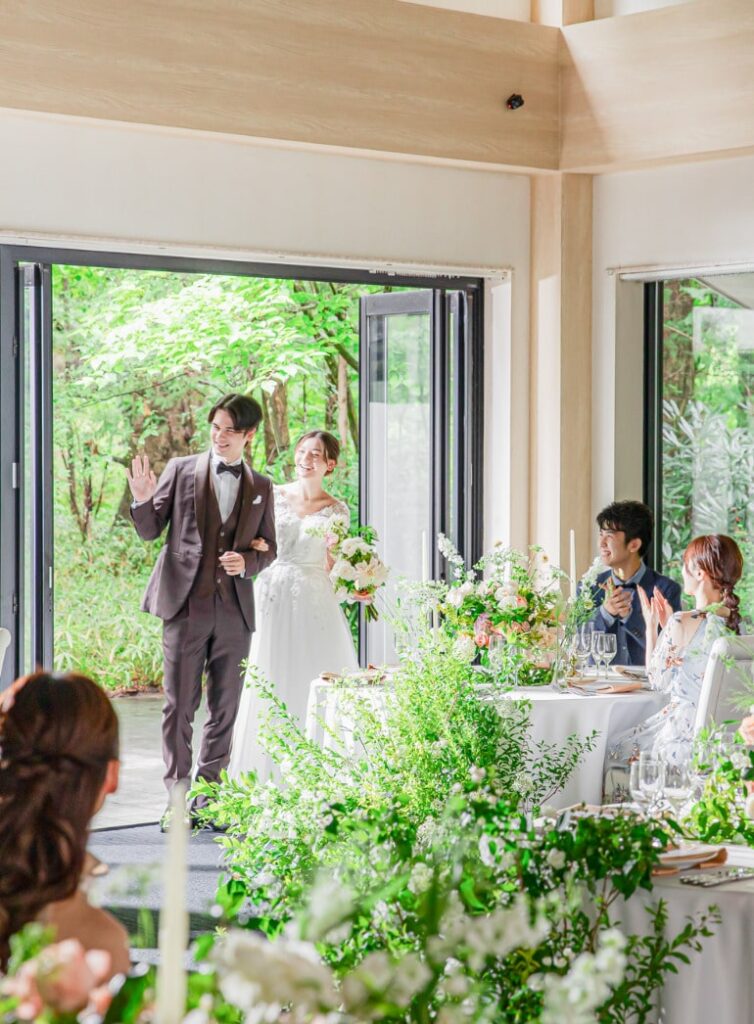アネーリ軽井沢の結婚式披露宴。新郎新婦が軽井沢の豊かな緑が望める開放的なテラスから入場する感動的なシーン。ウェディングドレスとブラウンのタキシード。テーブルには白とグリーンのナチュラルな装花が飾られ、ゲストハウスならではのアットホームな雰囲気。森の中のリゾートウェディングを求めるカップルに。