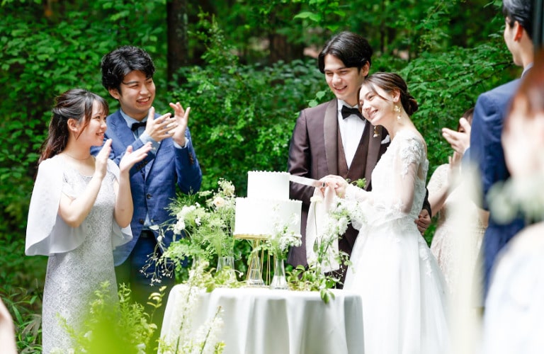 アネーリ軽井沢の結婚式。軽井沢の豊かな緑に囲まれたガーデンで、新郎新婦がウェディングケーキに入刀するセレモニー。純白のドレスとブラウンのタキシードが自然に映える。ゲストが温かく見守り、拍手で祝福するガーデンウェディングの様子。