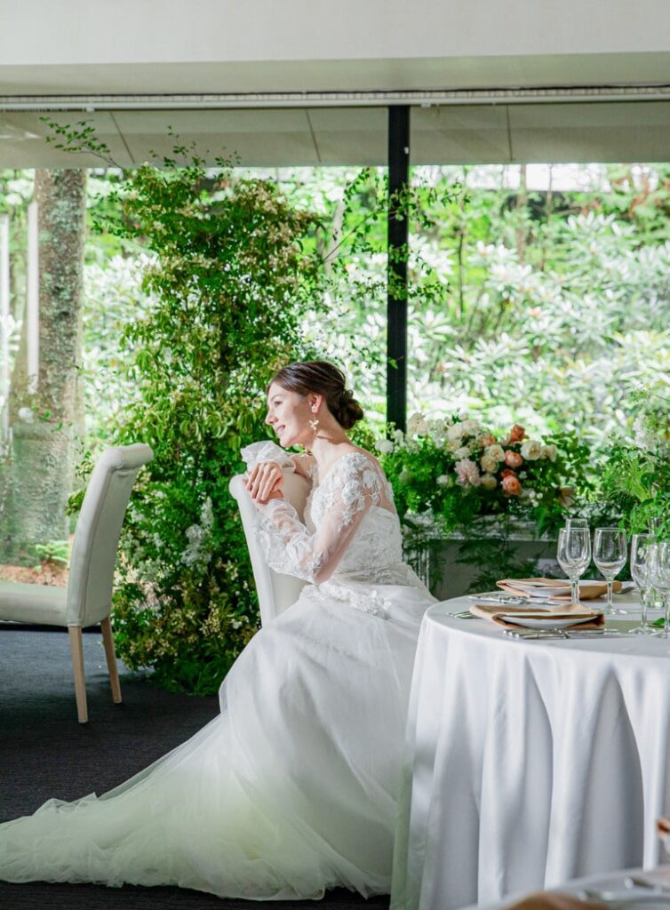 アネーリ軽井沢の披露宴会場で、窓外の軽井沢の緑を背景に座る新婦のポートレート。ウェディングドレスの繊細なレース袖が美しい。会場内は白とグリーンの装花でナチュラルに装飾され、リゾートウェディングらしい穏やかで開放的な雰囲気。