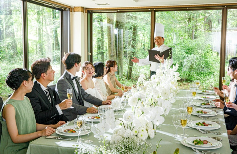 窓外の軽井沢の豊かな森を背景に、シェフが料理を紹介するシーン。新郎新婦とゲストが長テーブルを囲み、料理と歓談を楽しむゲストハウスウェディングの様子。白と緑の装花で飾られたテーブルと、温かな自然光が、リゾート婚らしいアットホームな雰囲気を演出。