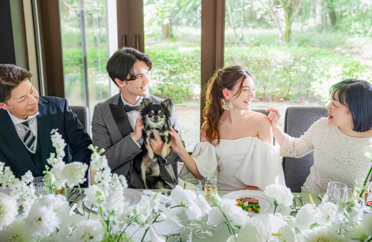 新郎新婦が愛犬と親族に囲まれ、和やかに過ごす披露宴。窓外の軽井沢の自然光が差し込み、白と緑の装花が飾られたテーブルが温かい雰囲気を演出。家族婚や親しい友人とのリゾート婚に最適なゲストハウス。