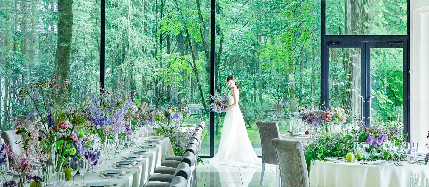 軽井沢の森の緑を背景にした、明るい披露宴会場。ゲストテーブルにはロングテーブルと円卓を配置。窓辺に立つ新婦のウェディングドレスが映える、ナチュラルリゾートウェディング。
