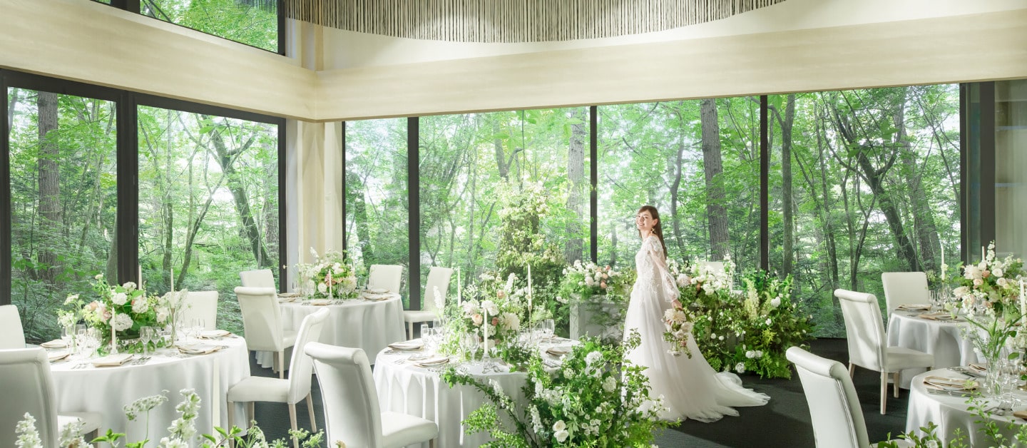 アネーリ軽井沢の結婚式披露宴会場。大きな窓の外に軽井沢の緑豊かな森が広がる、開放的な空間。白を基調としたテーブルコーディネートと、白とグリーンのナチュラルな装花。ウェディングドレス姿の新婦が森の中で佇むような、リゾートウェディングらしいロケーション。