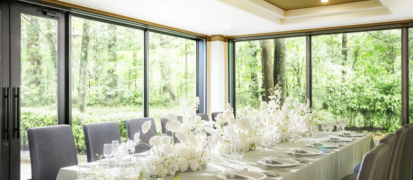 大きな窓から軽井沢の緑が望める開放的な空間。長テーブルに白の装花が豪華に飾られたテーブルコーディネート。シックな大人向けのリゾートウェディング、ゲストハウスの少人数婚や家族婚に最適なプライベートな雰囲気。