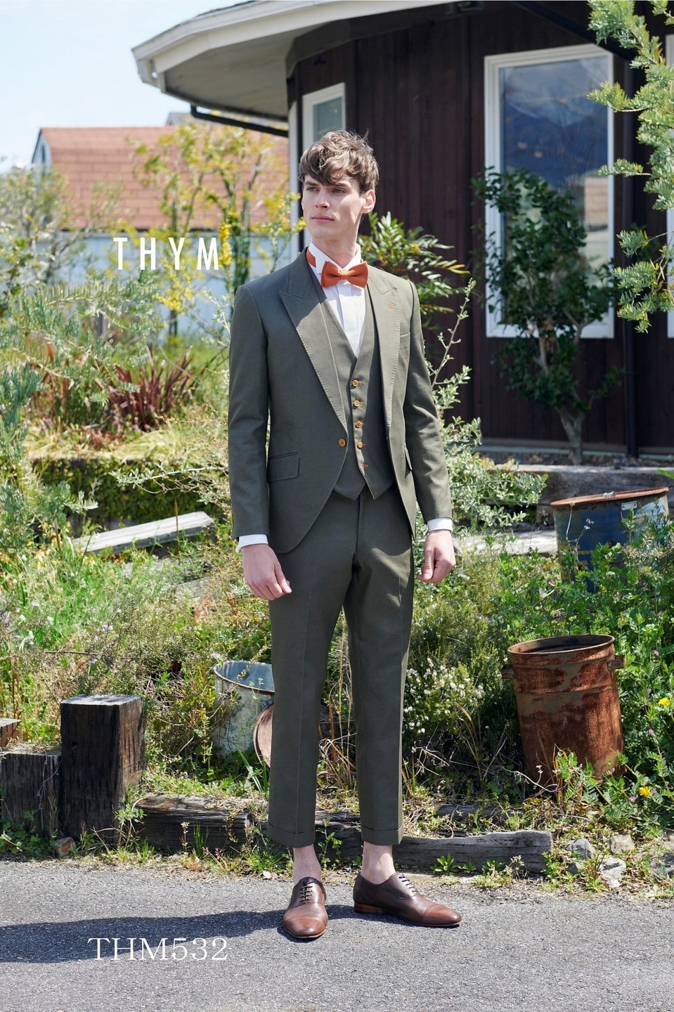 アネーリ軽井沢提携衣装。THYME (タイム) のカーキグリーンのタキシード (品番: THM532)。スリーピースでカジュアルシックに着こなせる新郎衣装。軽井沢の豊かな緑やガーデンウェディングでの前撮り・本番に最適なアースカラー。オレンジの蝶ネクタイがお洒落な、トレンド感溢れる1着。