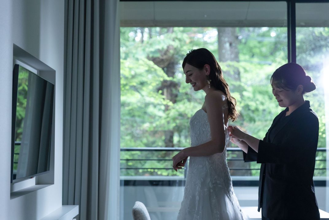 アネーリ軽井沢でのお支度シーン。提携宿泊施設の一室（ブライズルーム）で、ウェディングドレスを着付けてもらう花嫁。大きな窓から軽井沢の豊かな緑が見え、リラックスした雰囲気。軽井沢リゾートウェディングの当日、リラックスして準備を進めることができるブライズルームでの感動的な一枚。