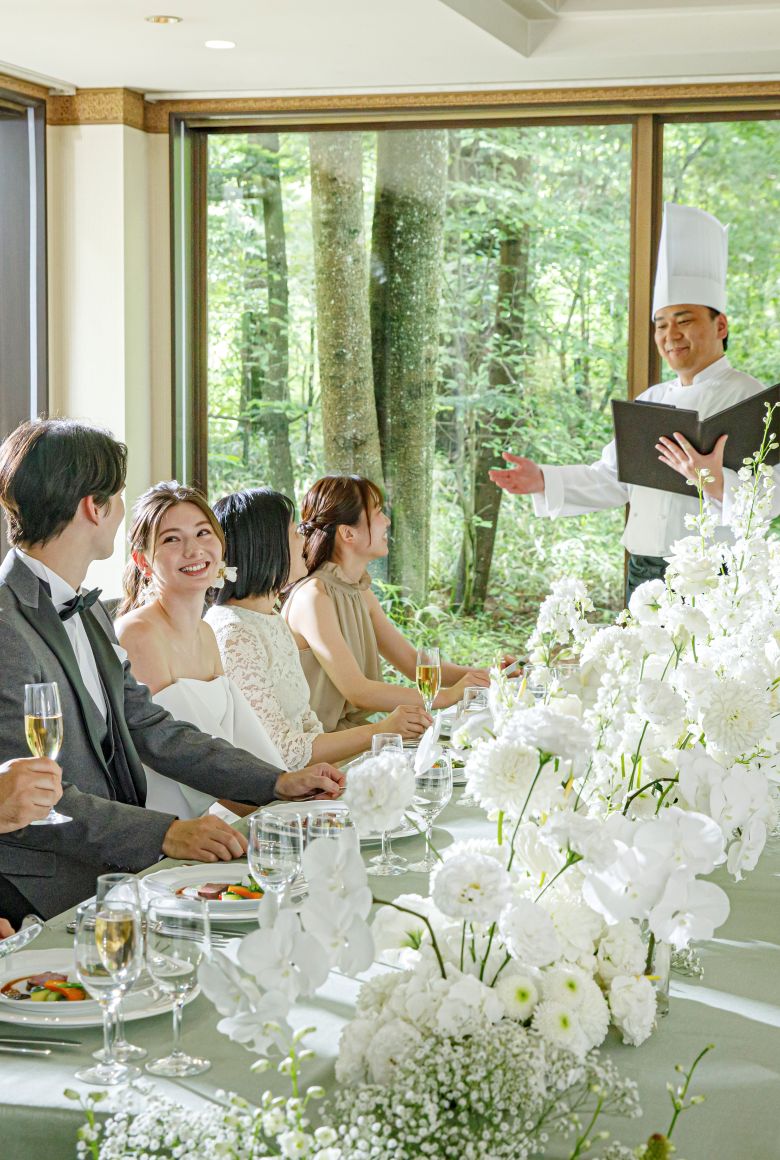 披露宴会場で、窓外の軽井沢の森の緑を眺めながら、新郎新婦とゲストが婚礼料理を楽しむシーン。シェフが直接料理説明をする演出で、ゲストへのおもてなしを重視。軽井沢の美食を楽しめるリゾートウェディングに最適な、開放的な会場での贅沢なひととき。