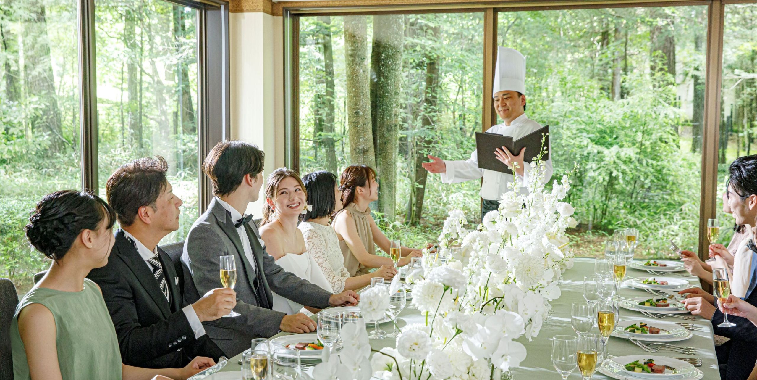 披露宴会場で、窓外の軽井沢の森の緑を眺めながら、新郎新婦とゲストが婚礼料理を楽しむシーン。シェフが直接料理説明をする演出で、ゲストへのおもてなしを重視。軽井沢の美食を楽しめるリゾートウェディングに最適な、開放的な会場での贅沢なひととき。