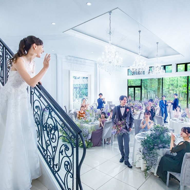 アネーリ軽井沢の白亜の大階段からの入場シーン。シャンデリアが輝く会場で、ゲストに手を振る花嫁と見守る新郎。憧れの階段演出が叶う披露宴。
