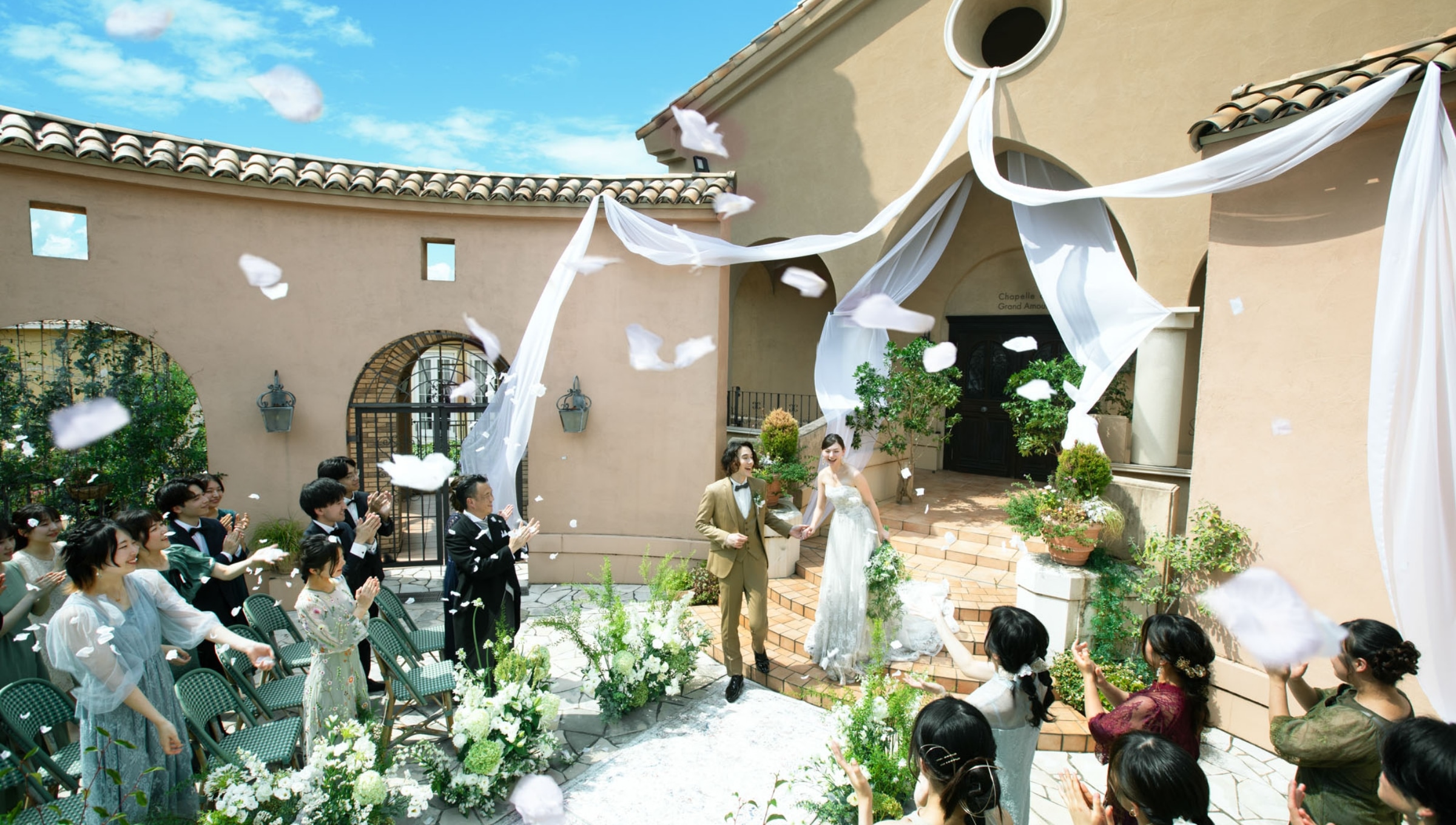 さいたま市の結婚式場アネーリ大宮。青空の下、白いドレープが飾られたガーデン。祝福のフラワーシャワーを浴びて、ゲストの歓声と拍手に包まれる新郎新婦。