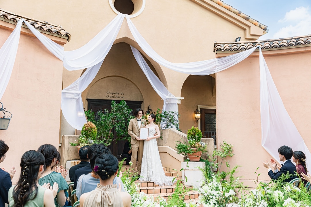 さいたま市の結婚式場アネーリ大宮。青空の下、白いドレープが飾られたガーデン挙式で、新郎新婦が結婚証明書を掲げ誓う感動的な瞬間