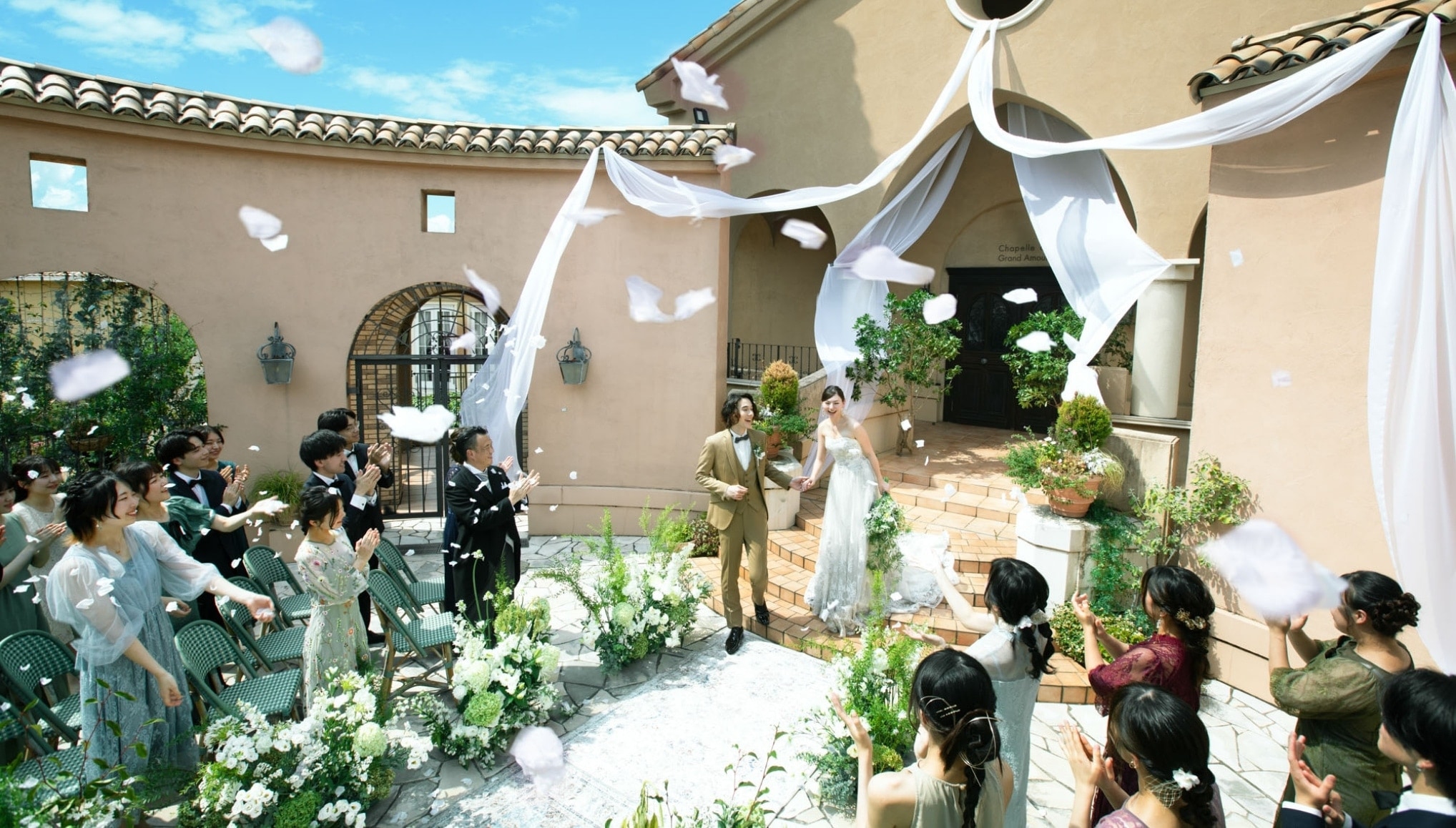 さいたま市の結婚式場アネーリ大宮。青空の下、ガーデンでフラワーシャワーの祝福を浴びる新郎新婦。ゲストの笑顔に包まれた開放的なアフターセレモニー。