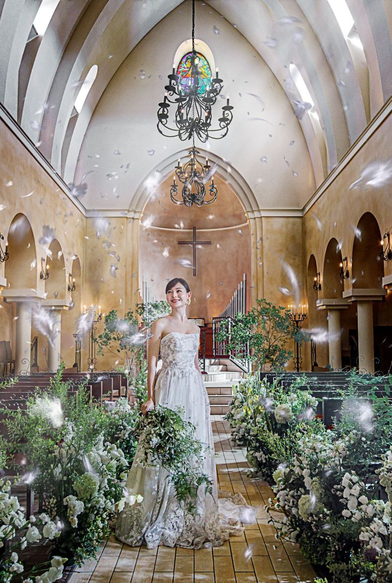 さいたま市の結婚式場アネーリ大宮。緑豊かな大聖堂に、天使の羽が舞い降りるフェザーシャワー。幻想的な光と祝福に包まれ、笑顔で佇む花嫁。