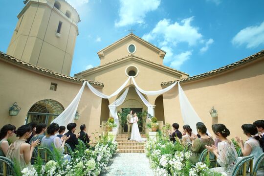 さいたま市の結婚式場アネーリ大宮。青空とチャペルを背景にした開放的なガーデン挙式。白いドレープと緑に囲まれ、ゲストの拍手に包まれる幸せな新郎新婦。