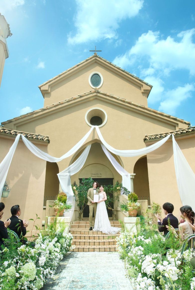 さいたま市の結婚式場アネーリ大宮。青空の下、白いドレープが揺れる中庭でのガーデン人前式。ゲストの祝福に包まれ、結婚証明書を掲げる新郎新婦。