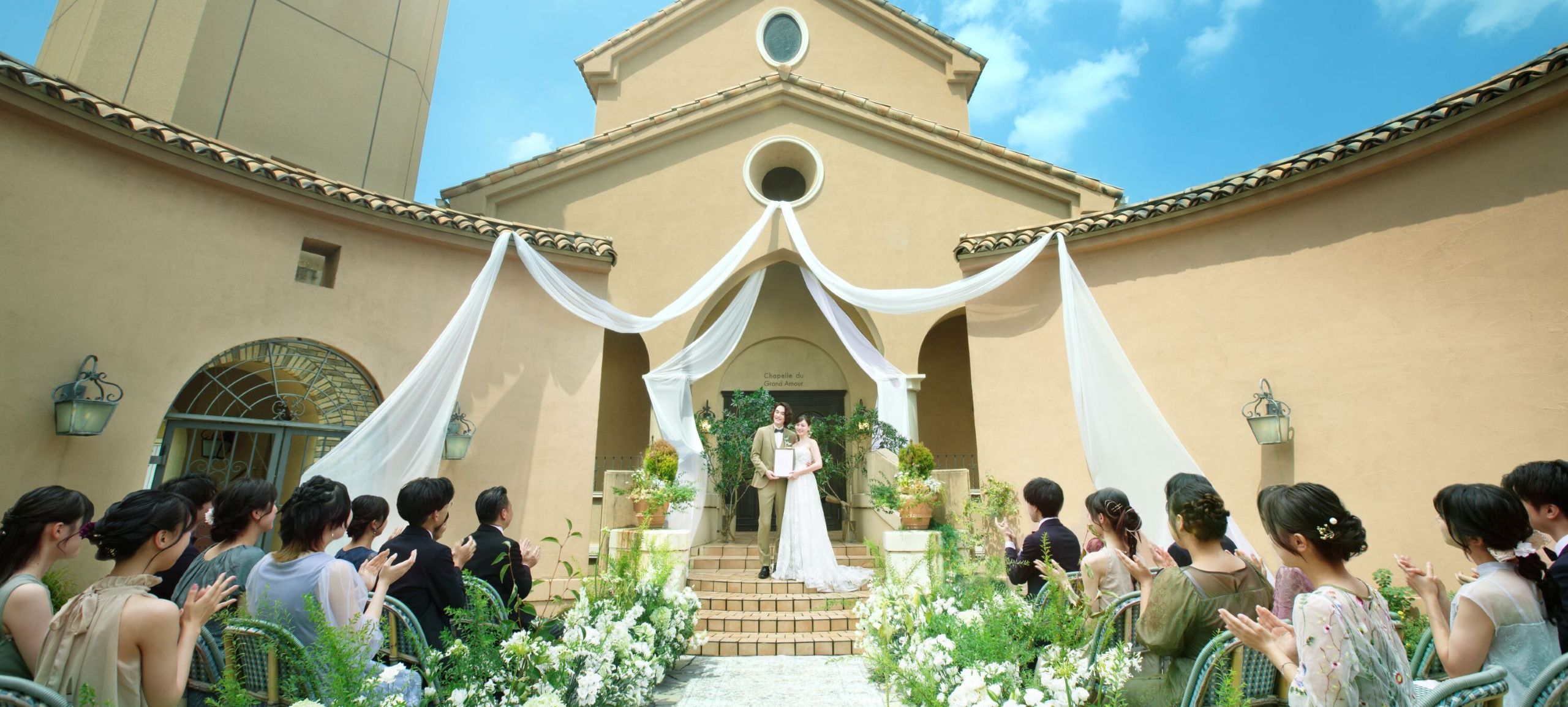 さいたま市の結婚式場アネーリ大宮。青空の下、白いドレープが揺れる中庭でのガーデン人前式。ゲストの祝福に包まれ、結婚証明書を掲げる新郎新婦。