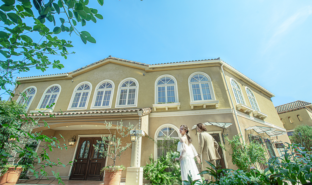 さいたま市の結婚式場アネーリ大宮。青空に映える南欧風邸宅と緑のガーデンを歩く新郎新婦。海外リゾートのような非日常感あふれるロケーションフォト。