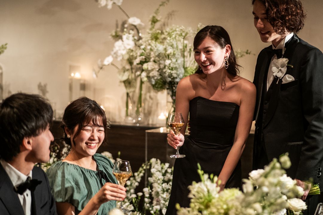 さいたま市の結婚式場アネーリ大宮。ブラックドレスの新婦と新郎が、ゲストのテーブルで笑顔で歓談。距離感が近いアットホームなナイトパーティ。