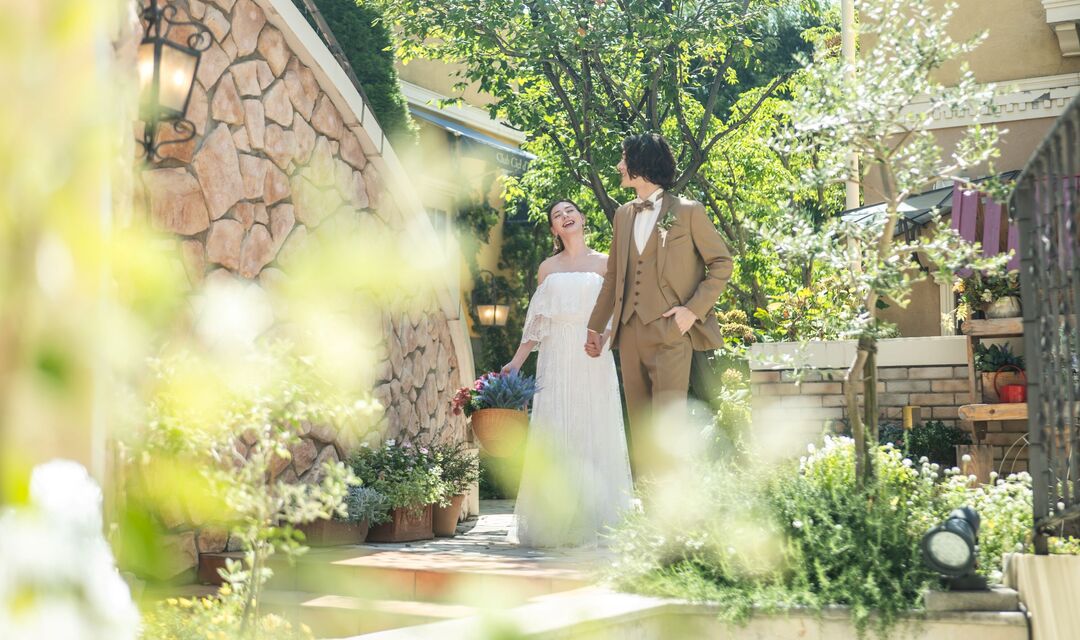 さいたま市の結婚式場アネーリ大宮。緑と光が溢れる南欧風のガーデンで、手を繋ぎ談笑しながら歩く新郎新婦。自然体な笑顔が輝く幸せなウェディングフォト。