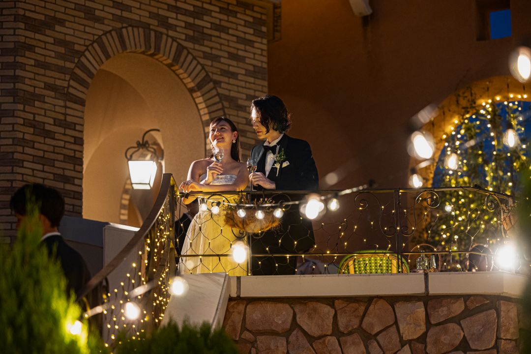 さいたま市の結婚式場アネーリ大宮。イルミネーションが輝くバルコニーで乾杯する新郎新婦。ゲストを見下ろし、ロマンチックなひとときを過ごす。