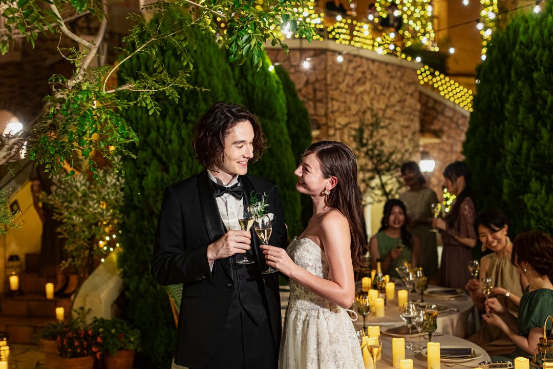 さいたま市の結婚式場アネーリ大宮。キャンドルの光に包まれたガーデンで、新郎新婦が笑顔で見つめ合い乾杯する。ロマンティックなナイトウェディング。