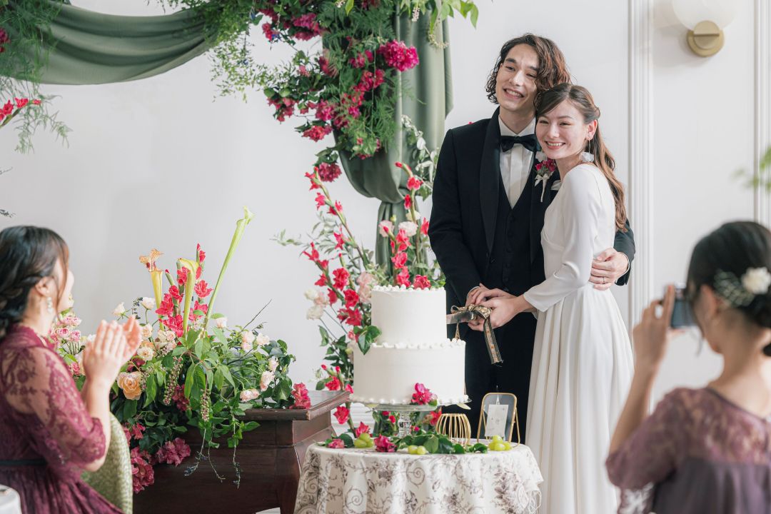 さいたま市の結婚式場アネーリ大宮。豪華な装花に囲まれ、新郎新婦がウェディングケーキ入刀。ゲストの拍手とカメラに包まれた、幸せな披露宴のワンシーン
