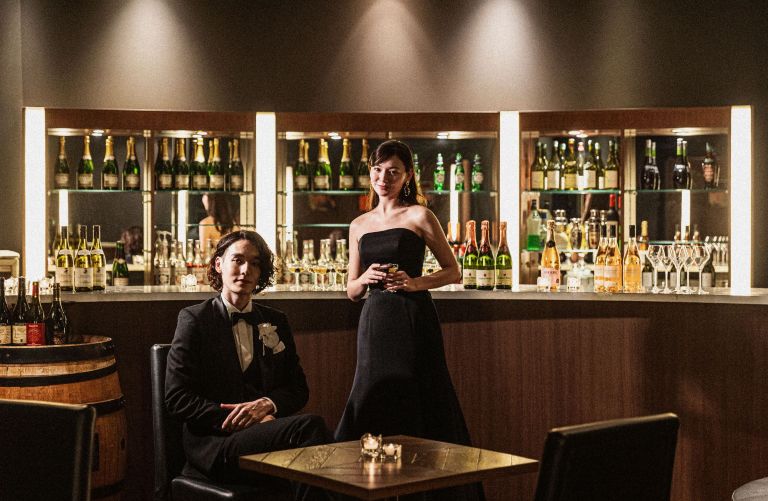 さいたま市の結婚式場アネーリ大宮。シックなバーカウンターでポーズをとる新郎新婦。ブラックドレスが映える、大人のナイトウェディングのフォトスポット。