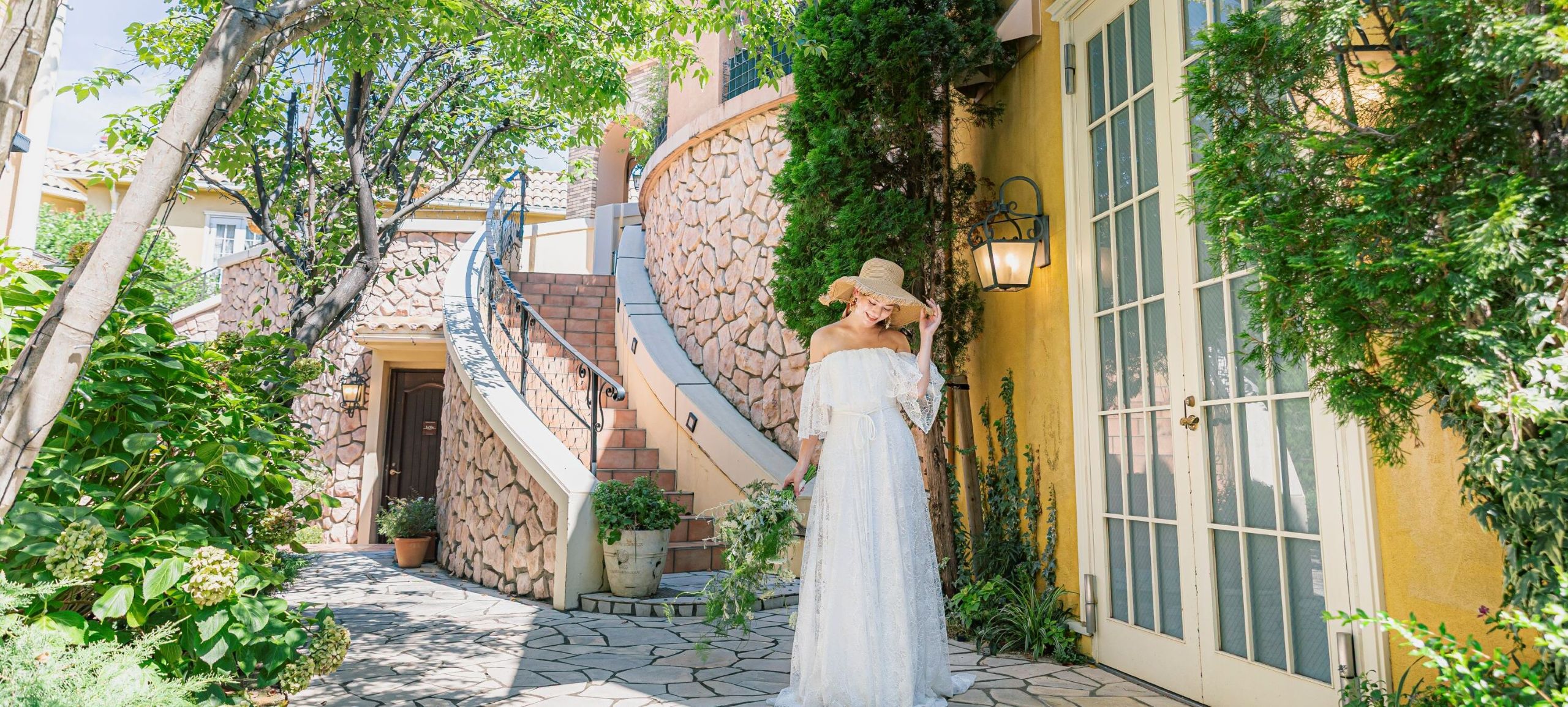 さいたま市の結婚式場アネーリ大宮。南欧風の螺旋階段と黄色い壁が続く街並みで、麦わら帽子の花嫁が佇む。まるで海外リゾートのようなナチュラルな一枚。