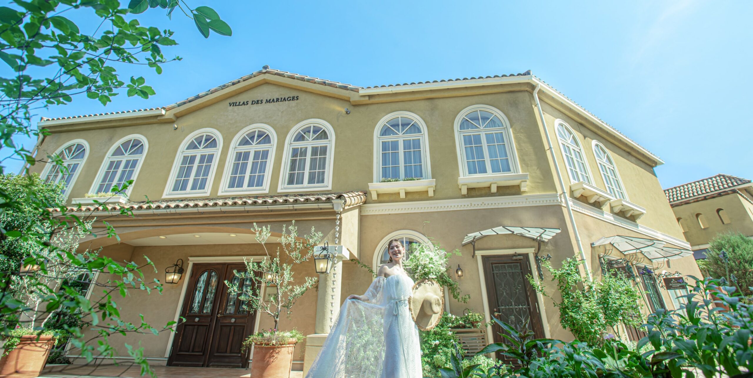 さいたま市の結婚式場アネーリ大宮。青空の下、南欧風の邸宅と緑に囲まれたガーデンで、ウェディングドレス姿の花嫁が微笑むリゾート感あふれる一枚