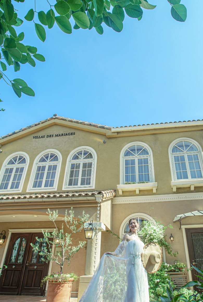 さいたま市の結婚式場アネーリ大宮。青空の下、南欧風の邸宅と緑に囲まれたガーデンで、ウェディングドレス姿の花嫁が微笑むリゾート感あふれる一枚