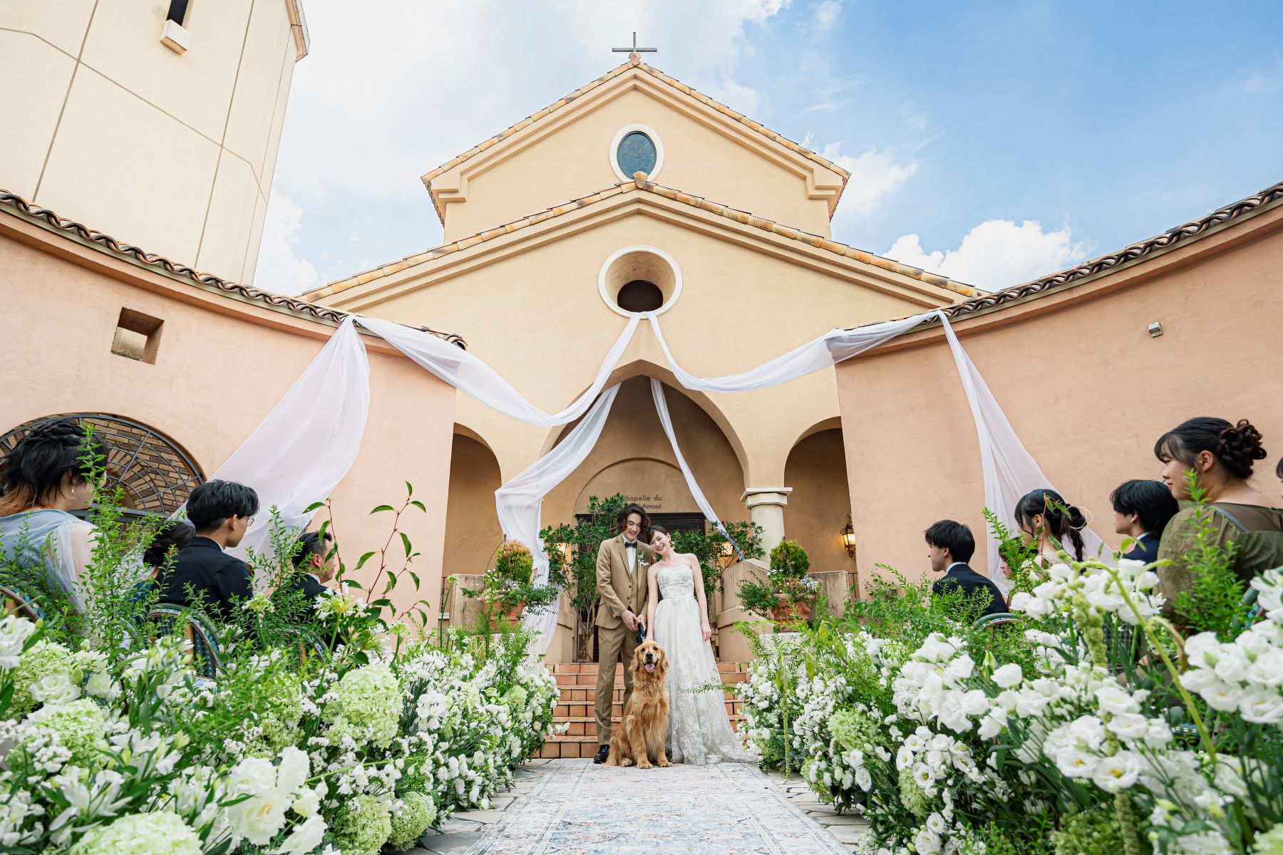 「大宮の結婚式場『アネーリ大宮』の広大なガーデンで行われる、愛犬も参加可能なガーデン人前式。青空が広がる屋外チャペルをバックに、大切なペットと一緒に誓いを立てる新郎新婦。豊かな緑とゲストの祝福に包まれた、開放感あふれるナチュラルなペットウェディングの様子。」