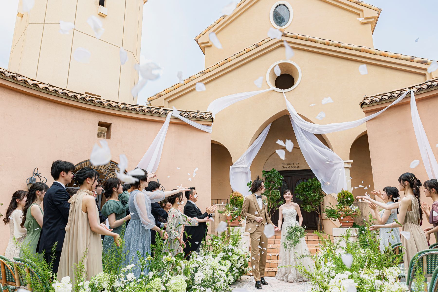 「大宮の結婚式場『アネーリ大宮』の開放的なガーデン人前式で行われるフラワーシャワー。壮麗な屋外チャペルを背景に、ゲストが舞い上がらせる花びらの中を笑顔で進む新郎新婦。祝福に包まれたアットホームでナチュラルなウェディングの感動的なワンシーン。」