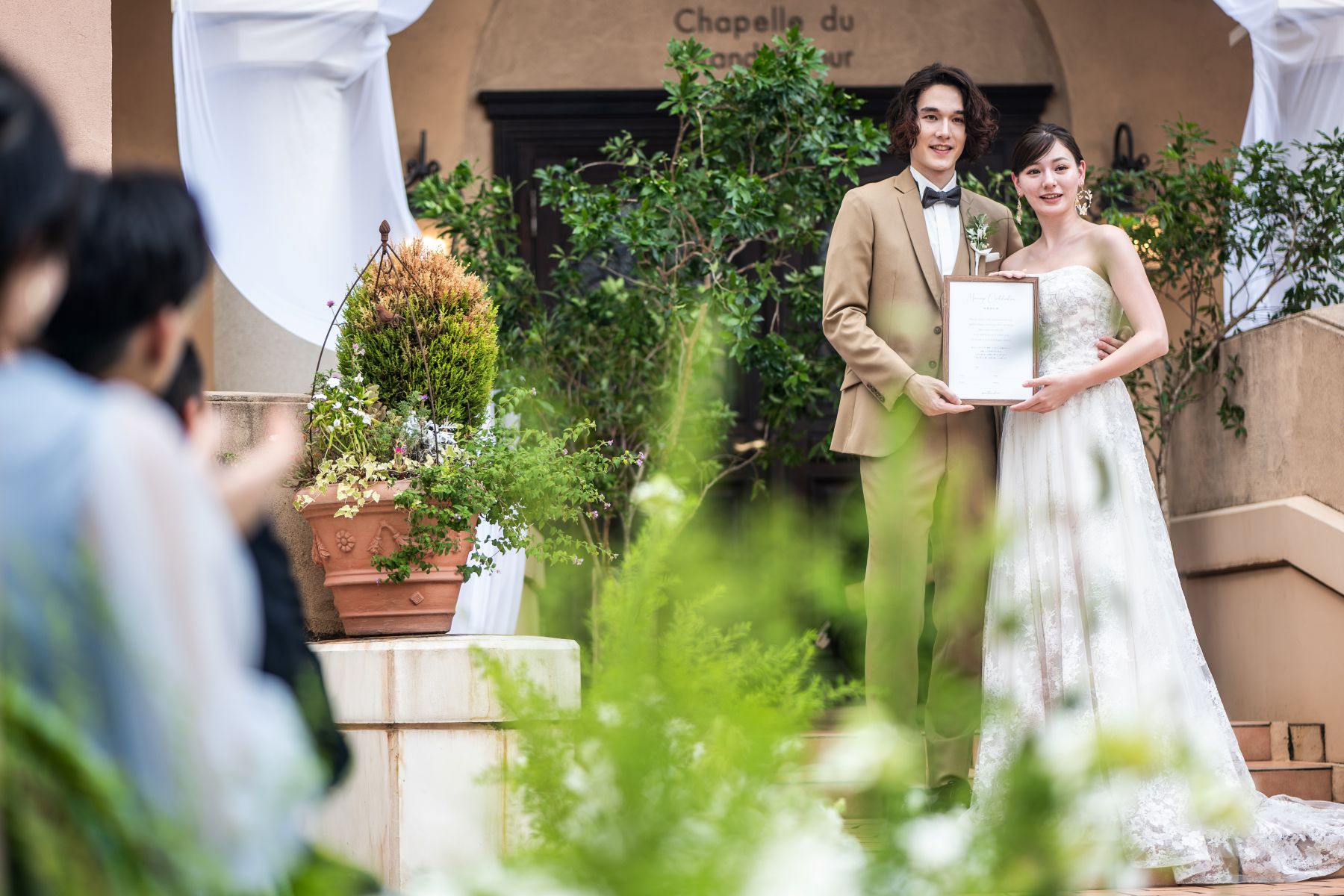 「大宮の結婚式場『アネーリ大宮』の開放的なガーデンで行われる人前式の様子。緑豊かな屋外チャペルを背景に、タキシードとウェディングドレス姿の新郎新婦が笑顔で結婚証明書を披露する、アットホームでナチュラルなウェディングシーン。」