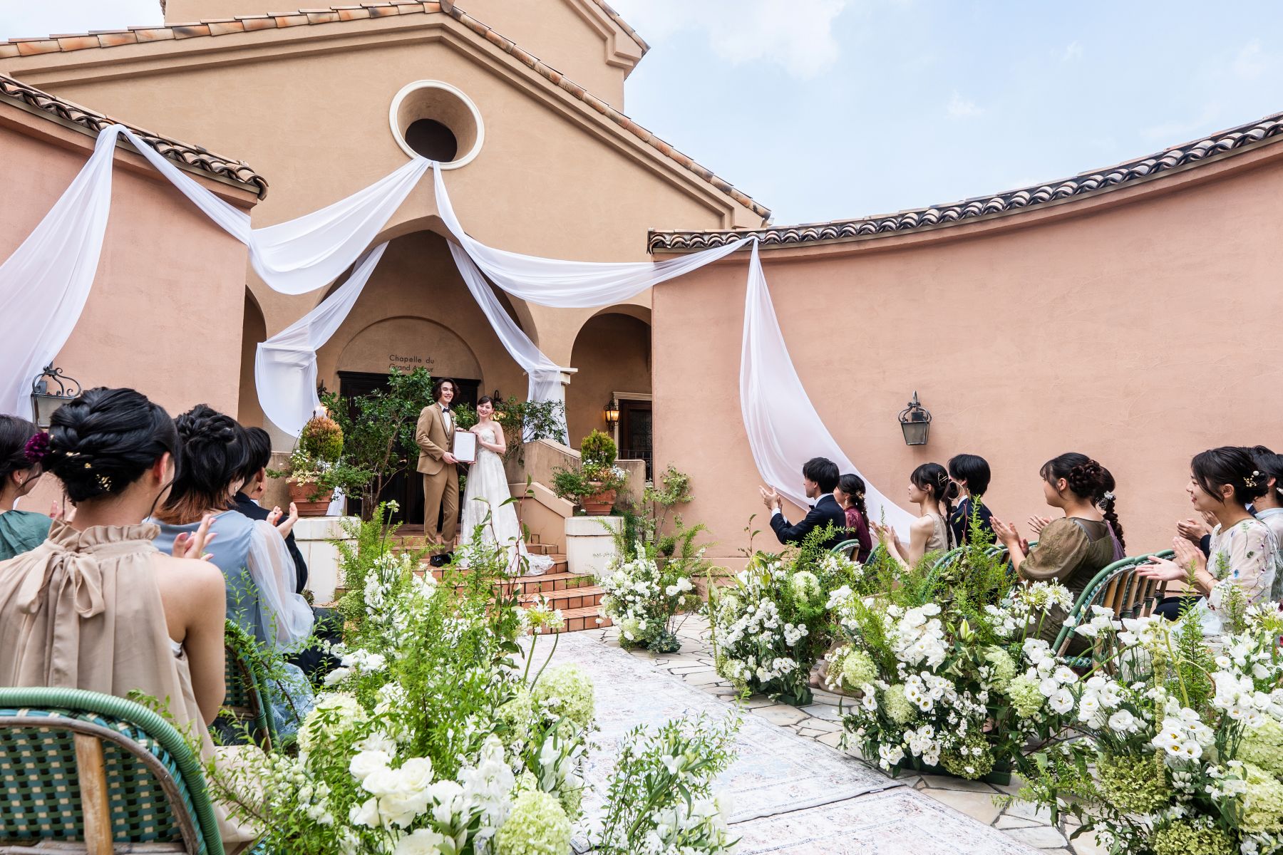 「大宮の結婚式場『アネーリ大宮』の開放的なガーデン人前式。緑豊かな屋外チャペルを背景に、ゲストの温かな拍手に包まれながら新郎新婦が笑顔で結婚証明書を披露する、アットホームでナチュラルなウェディングシーン。」