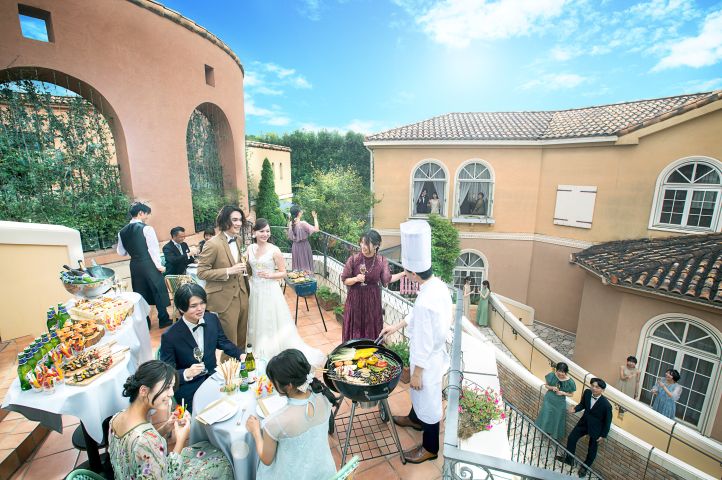 アネーリ大宮の青空広がるテラスで、シェフが振る舞うBBQ演出を楽しむ新郎新婦とゲスト。さいたま市の欧風邸宅を貸し切って叶える、アットホームで開放的なガーデンウェディングのパーティーシーン。