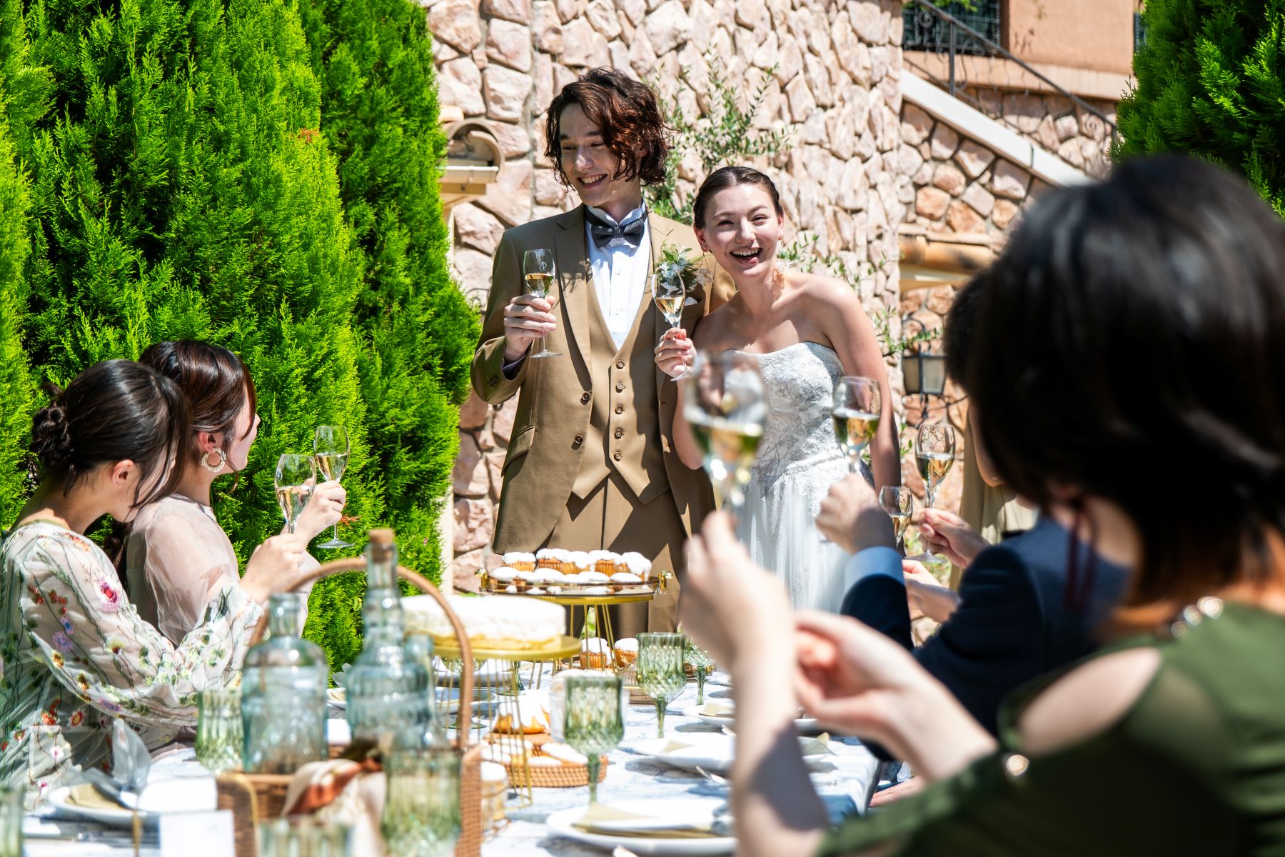 さいたま市の結婚式場「アネーリ大宮」の屋外テラスでのガーデンウェディング。豊かな緑と温かみのある石壁を背景に、新郎新婦とゲストが笑顔で乾杯を交わすアットホームなパーティーの様子。自然光が降り注ぐ開放的な空間での屋外演出シーン。