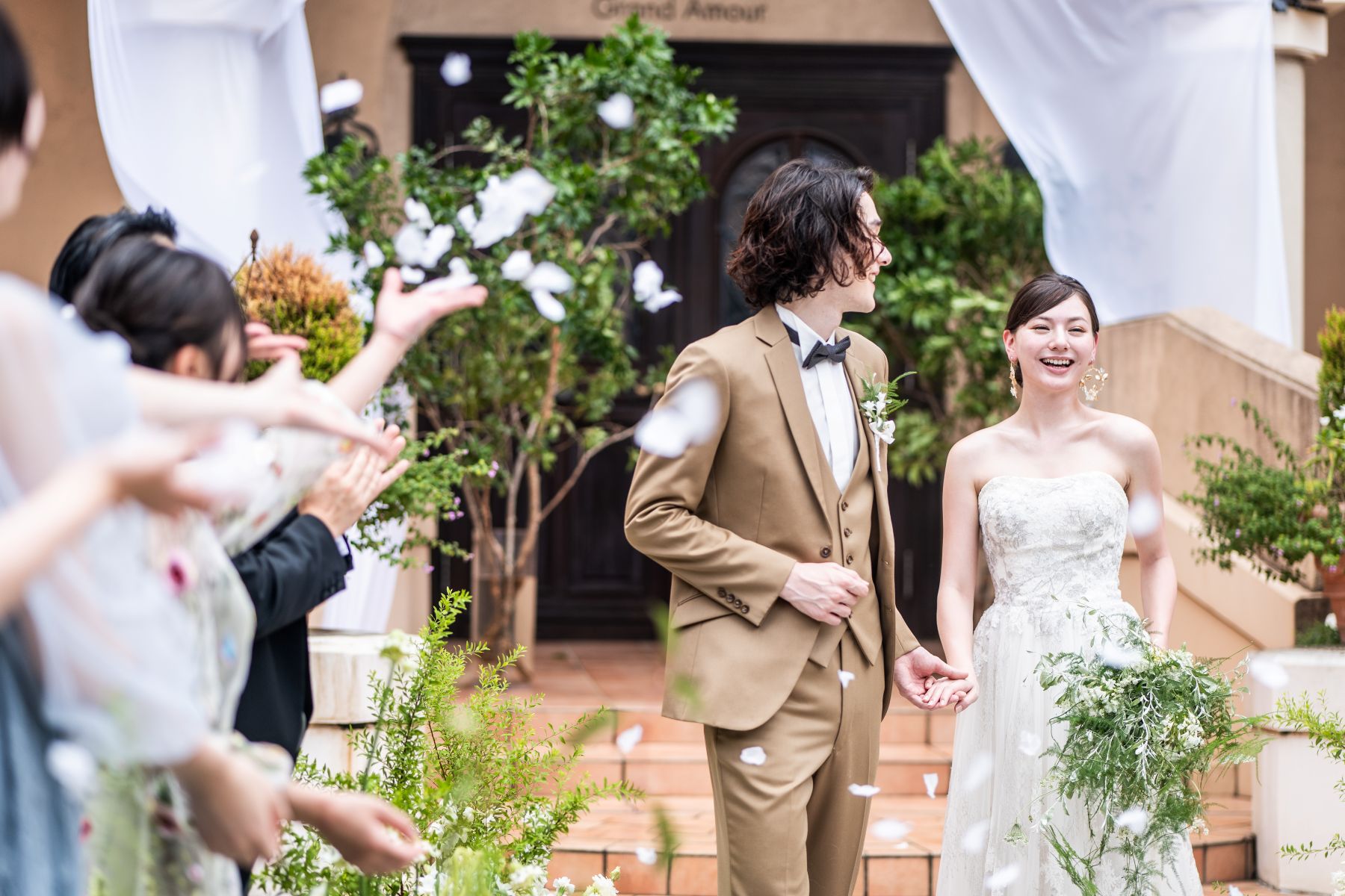 「大宮の結婚式場『アネーリ大宮』のガーデン人前式。挙式後のフラワーシャワーで、ゲストからの温かな祝福と花びらに包まれ、満面の笑みで進む新郎新婦。開放感あふれる屋外チャペルでの、ナチュラルでアットホームなウェディングのワンシーン。」